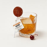 Old Fashioned Bourbon Lollipop with its Double Neat hang tag, leaning on an old fashioned cocktail